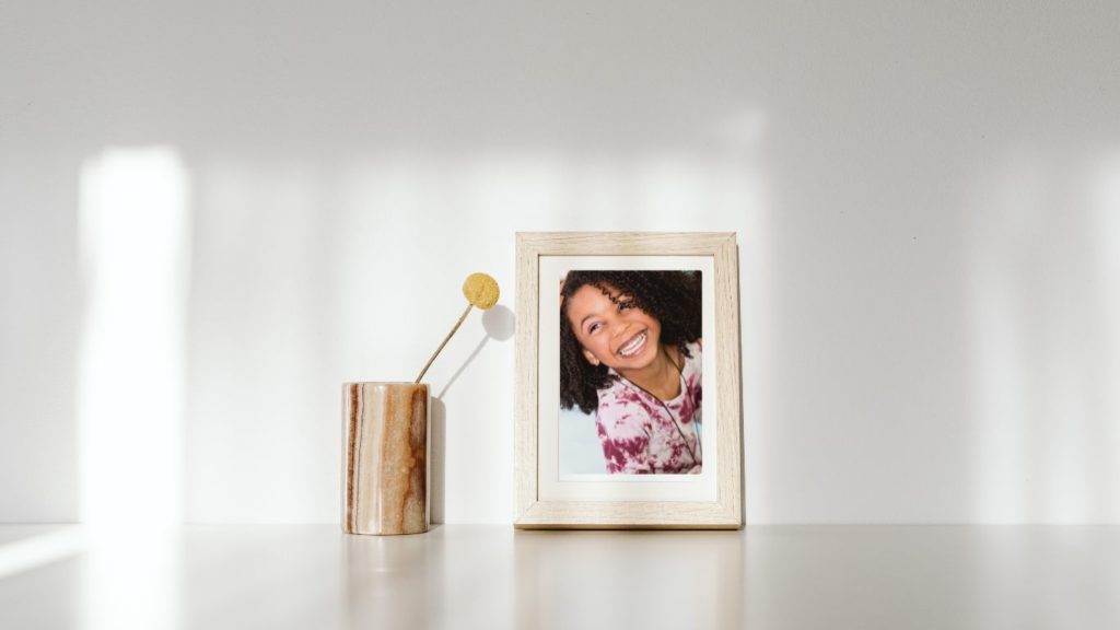 framed picture of an African American child with curly hair sitting on a desk with a brown and cream cylindrical vase sitting beside it that contains a single yellow pom flower as a window shadow is cast on the scene 