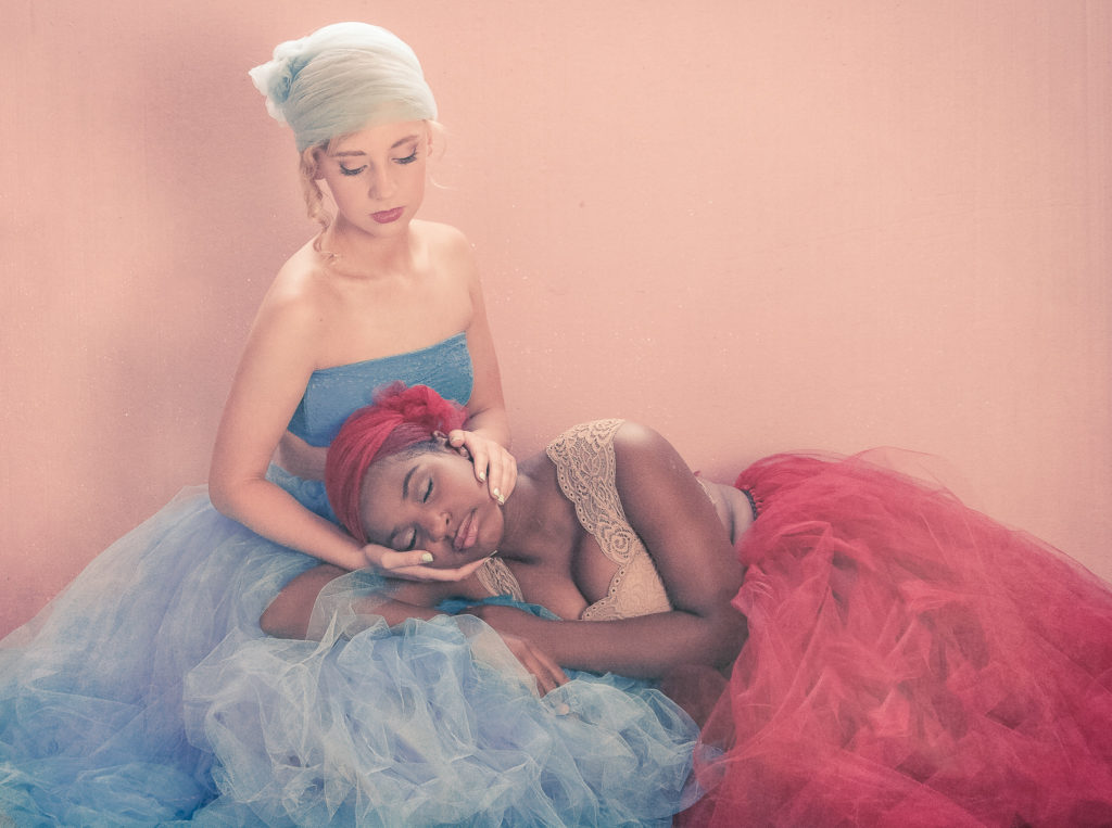 Caucasian girl with blonde hair  dressed in blue tulle caressing the face of an African American girl dressed in red tulle as she is lying with her eyes closed on the lap of the Caucasian girl captured by a Portrait photographer