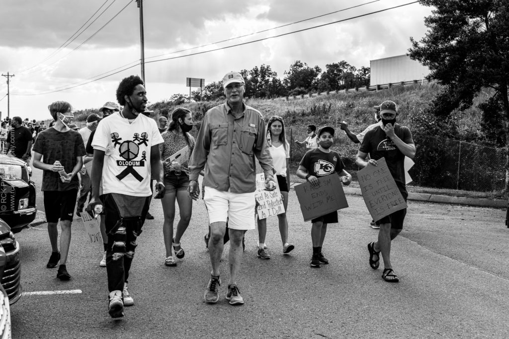 black and white image of a Black Man walking with several Caucasian protestors signs during a Black Lives Matter March. Captured by Denetra Smith of RCK Photography