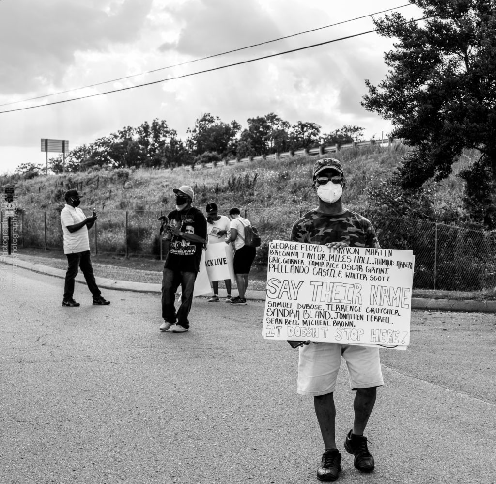 black and white image of a Caucasian man Holding a sign with "Say Their Name" and all the names of  Black victims of police brutality written on the sign. Captured by  Denetra Smith of RCK Photography