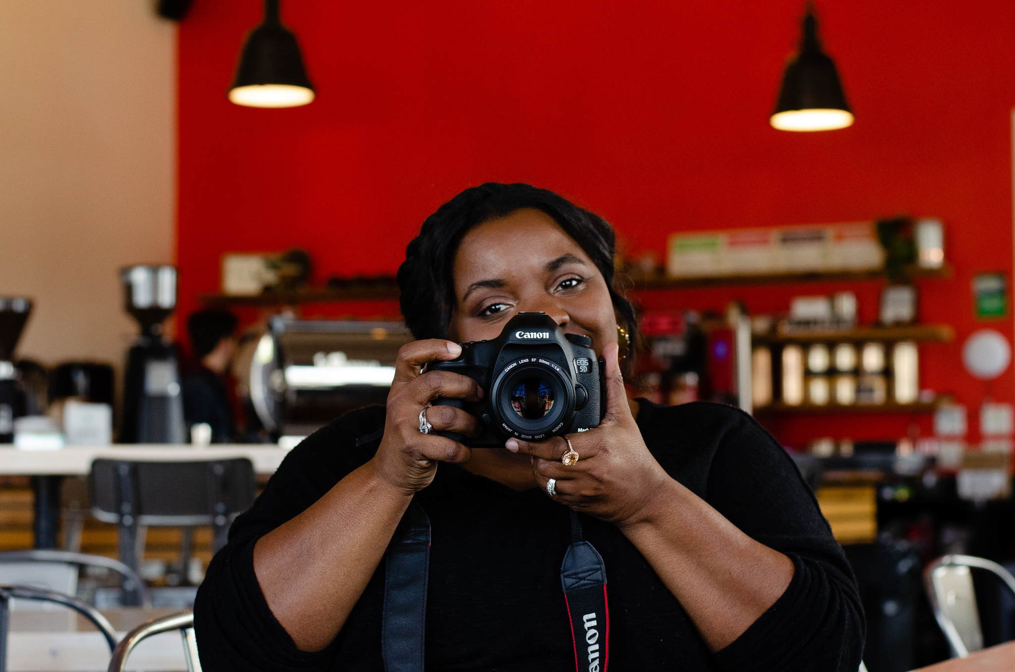 African American Female wearing aa black shirt holding a Canon camera in front of a red background at a coffee shop