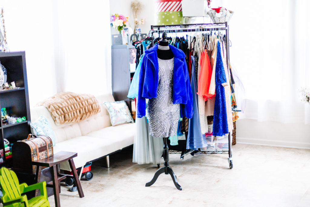 A Natural light studio featuring a wardrobe rack full of clothes, a white leather futon, props, and a mannequin wearing a silver dress diamond choker and royal blue fur coat captured by RCK Portrait Photographer Denetra Smith